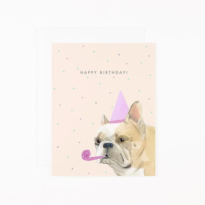 A birthday card featuring a watercolor illustration of a French bulldog wearing a pink party hat and blowing a pink party blower, with the text 'HAPPY BIRTHDAY!' on a pale pink background with small scattered confetti dots. A white envelope is partially visible behind the card.