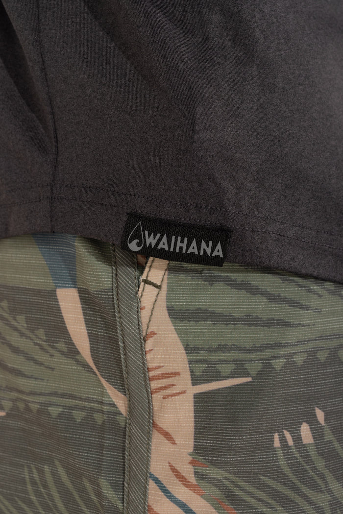 Close-up of the Waihana woven logo label on the hem of a charcoal Mariner hooded long sleeve sun shirt, layered over tropical-print shorts.