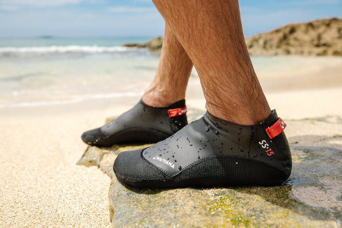 Close-up of a man's feet wearing wet black Waihana SS.15 neoprene dive booties with red pull tabs, standing on a mossy rock at a sandy beach with turquoise ocean in the background.