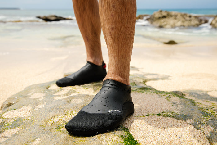 Close-up of a man's feet wearing black Waihana neoprene dive socks with water droplets on them, standing on a mossy rock with a sandy beach and turquoise ocean in the background.