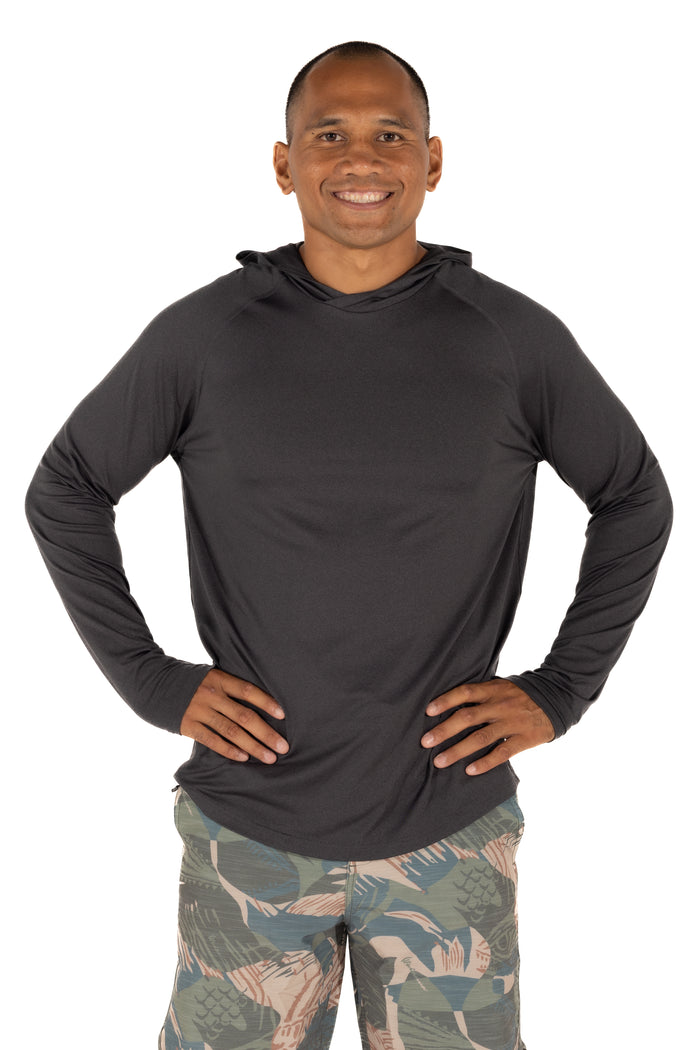 Male model wearing the Waihana Mariner Black Hooded Long Sleeve sun shirt in charcoal, paired with tropical-print board shorts, hands on hips, white studio background.