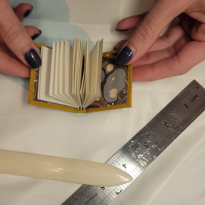 Hands holding small book with bone folder and ruler on a colorful surface