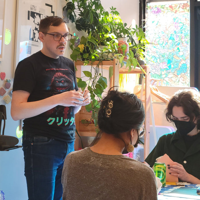 Person standing with two people sitting at a table with art supplies and a plant in the background.