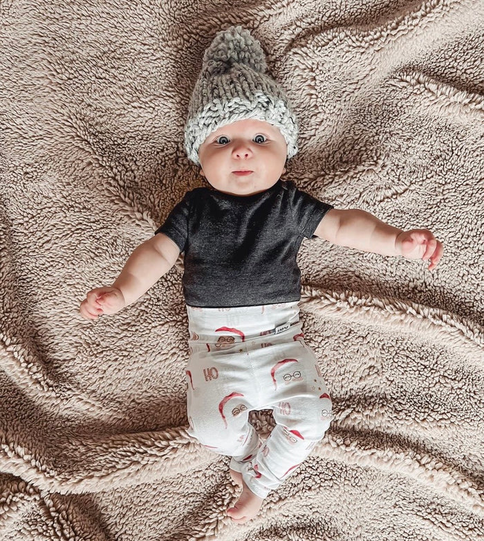 Baby wearing Vintage Santa leggings with festive red and white print, styled with a black top and a cozy gray knit hat, lying on a soft beige blanket. Perfect holiday outfit for babies and toddlers.
