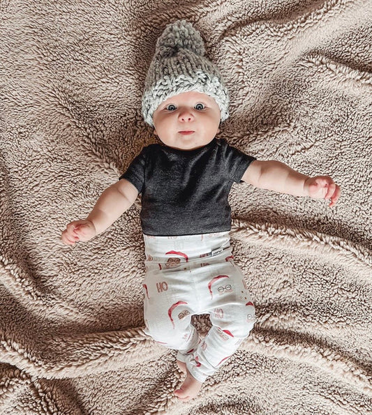 Baby wearing Vintage Santa leggings with festive red and white print, styled with a black top and a cozy gray knit hat, lying on a soft beige blanket. Perfect holiday outfit for babies and toddlers.